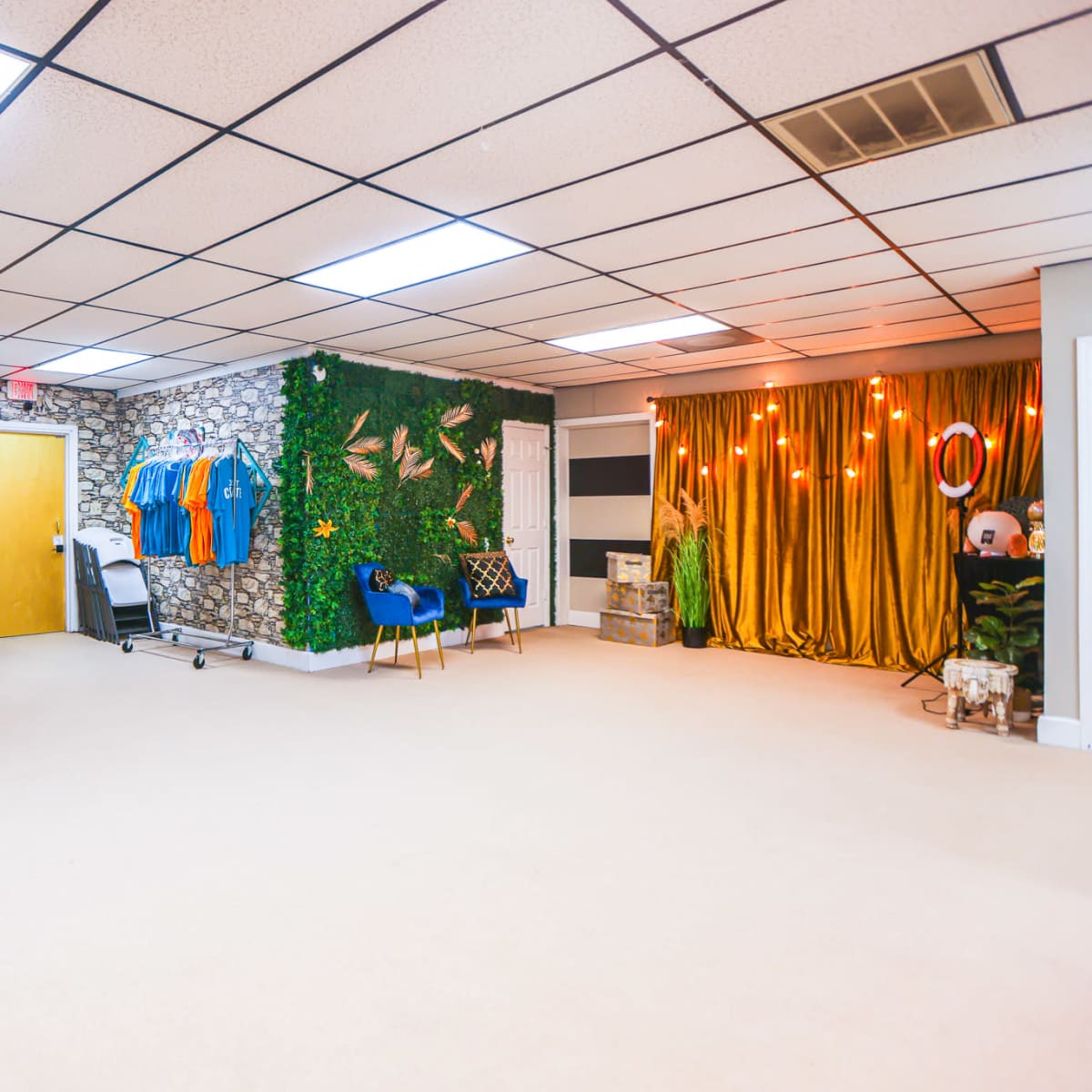 A spacious room features a stone wall adorned with clothing, a golden curtain backdrop with lights, and green foliage decor.