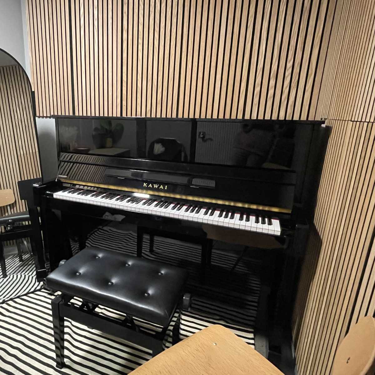 A Kawai piano in a room with vertical wooden paneling, accompanied by a mirrored wall and a small seating area.