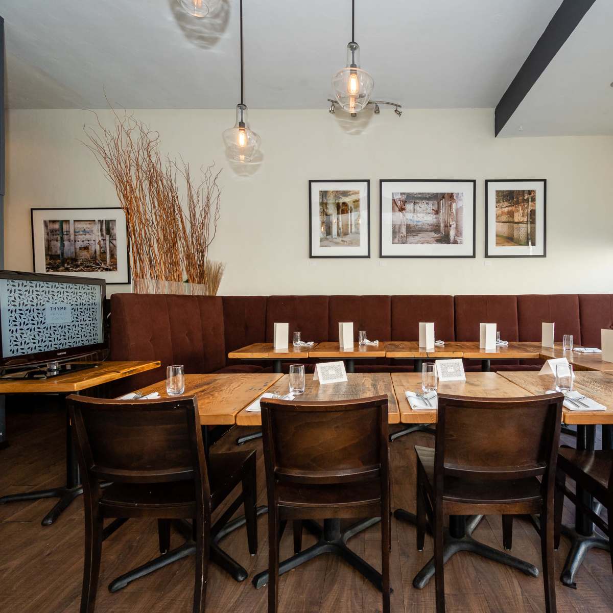 A dining area features a long, wooden table set for a meal, surrounded by brown chairs, with framed photographs and decorative plants in the background.