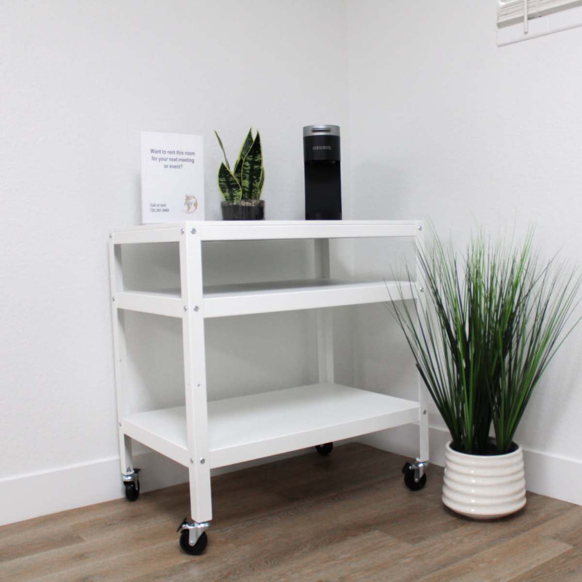 A white rolling cart with two shelves is positioned in a corner next to a coffee maker and a tall potted plant.