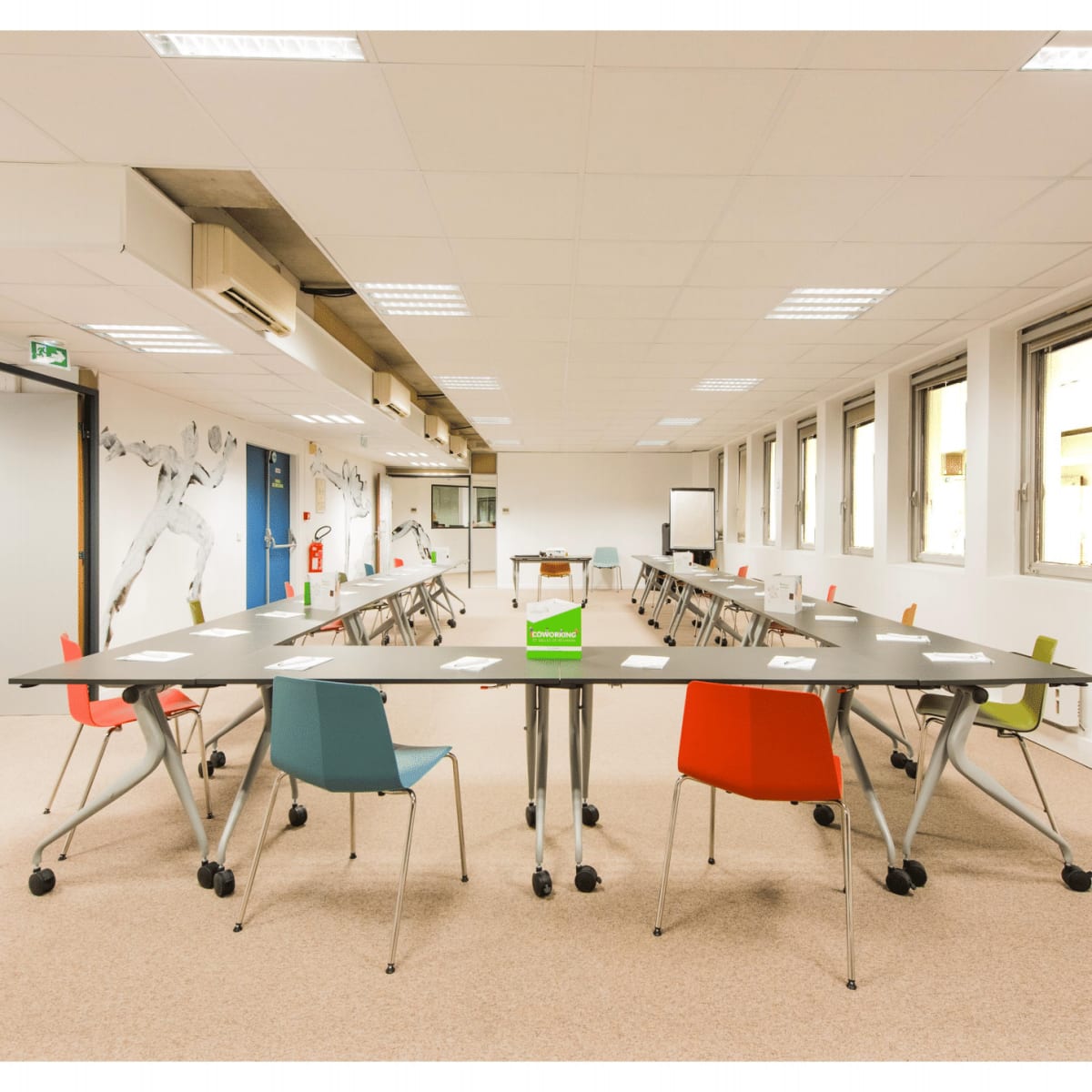 A modern meeting room features a circular arrangement of tables and colorful chairs with large windows and a whiteboard at the front.