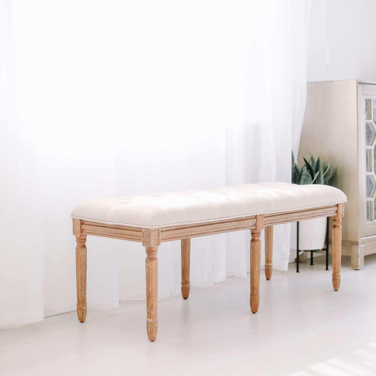 A wooden bench with a padded seat is positioned in a brightly lit room with sheer curtains.