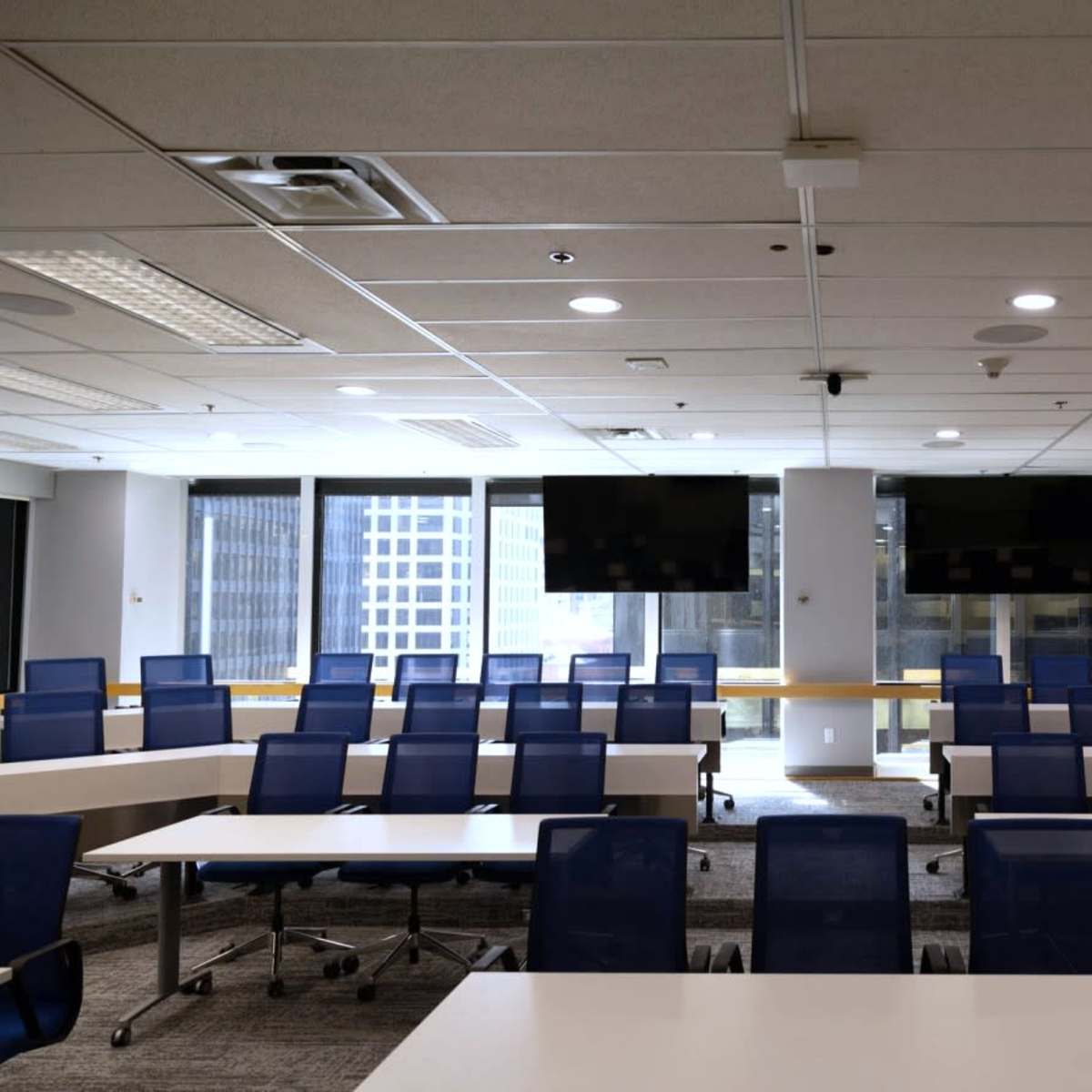 A modern conference room with rows of blue chairs and tables, large windows, and two flat-screen monitors on the wall.