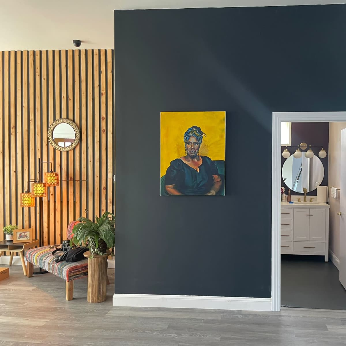A modern interior featuring a dark wall with a portrait, a stylish seating area with a plant, and an adjoining room visible through an open doorway leading to a bathroom.