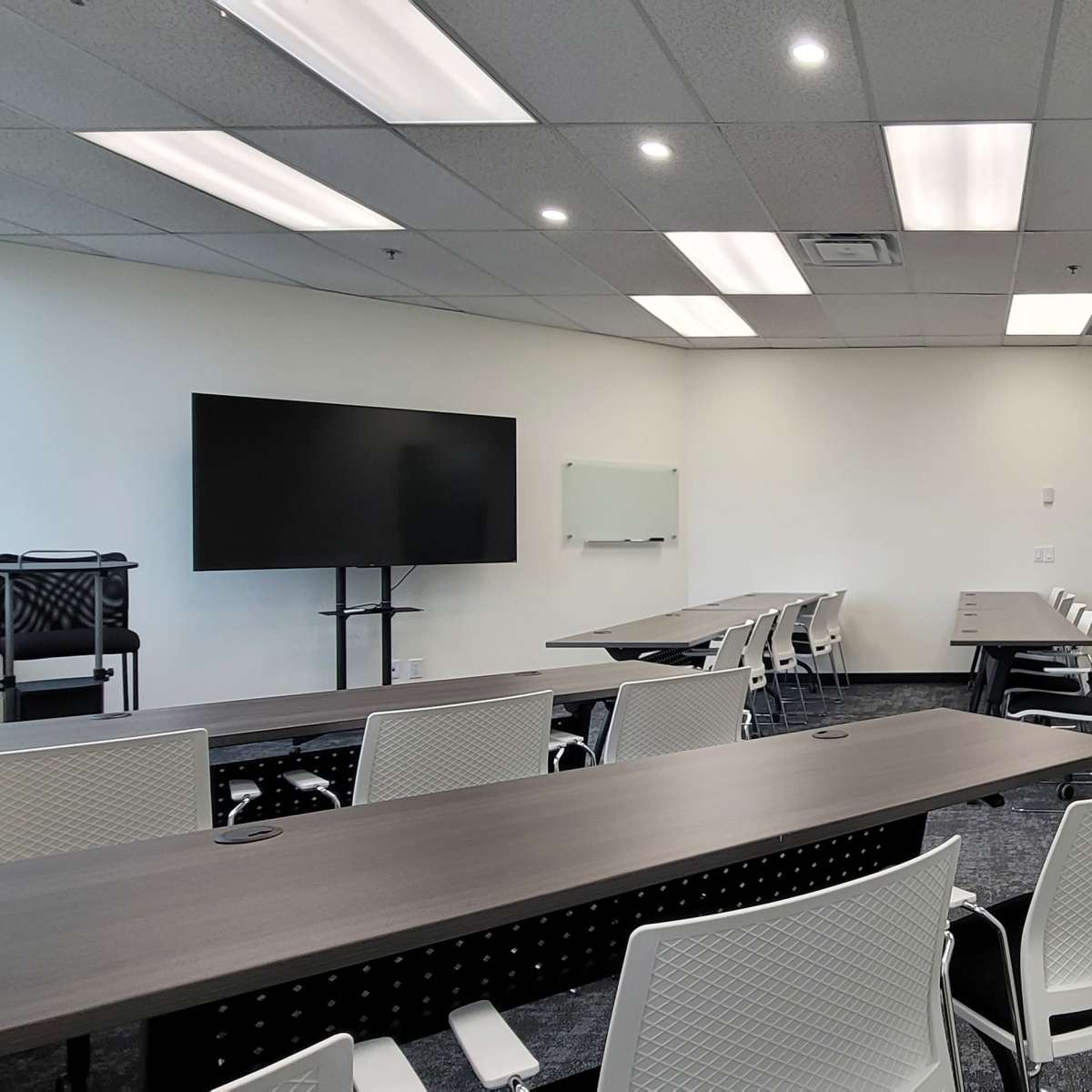 A meeting room with a large screen, whiteboard, and several white chairs arranged around a long table.