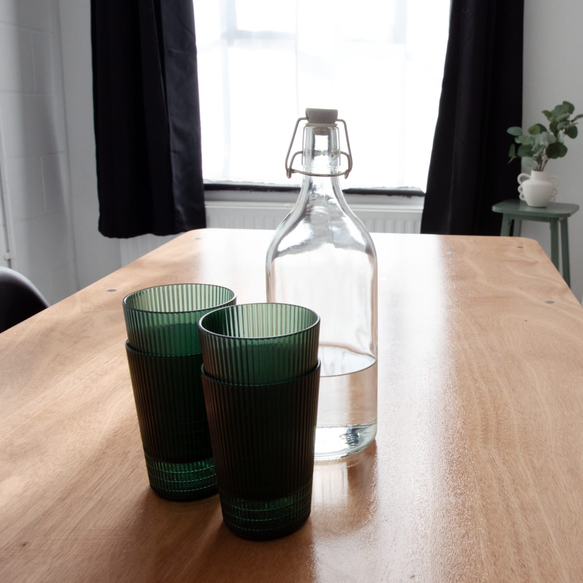A wooden table with two green glasses and a bottle of water sits in front of a cream-colored sofa and a potted plant in a well-lit room.