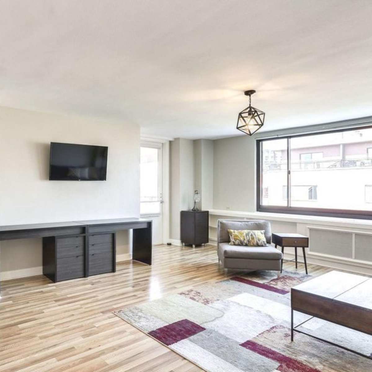 A modern living room with hardwood floors, a wall-mounted television, and a window providing natural light.