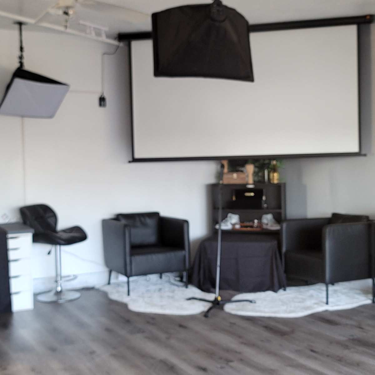 A well-equipped studio space featuring a large projection screen, multiple lighting setups, and various black furniture pieces arranged around a central table.