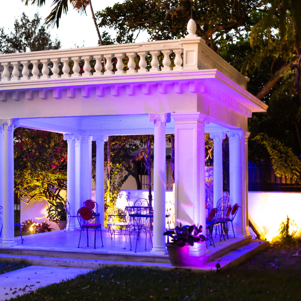 A white pavilion with columns and colorful lighting features seating arrangements in a landscaped garden.