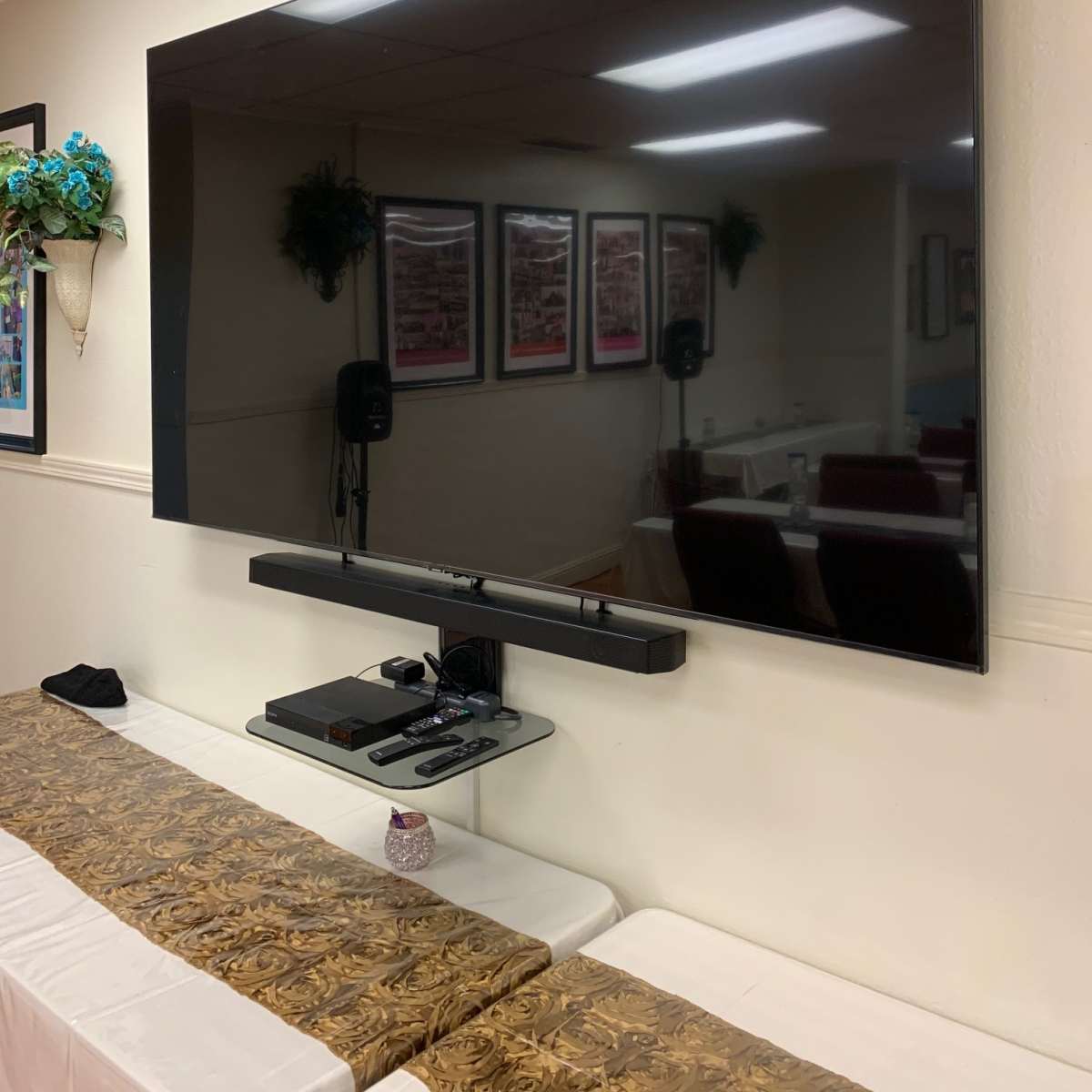 A large flat-screen TV is mounted on a wall above a soundbar and media device on a table in a room with framed pictures.