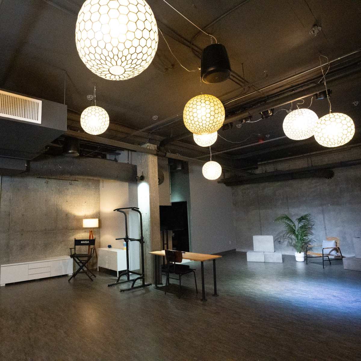 A spacious, modern room with concrete walls and several decorative pendant lights hanging from the ceiling, featuring a table, a chair, and a potted plant in the corner.