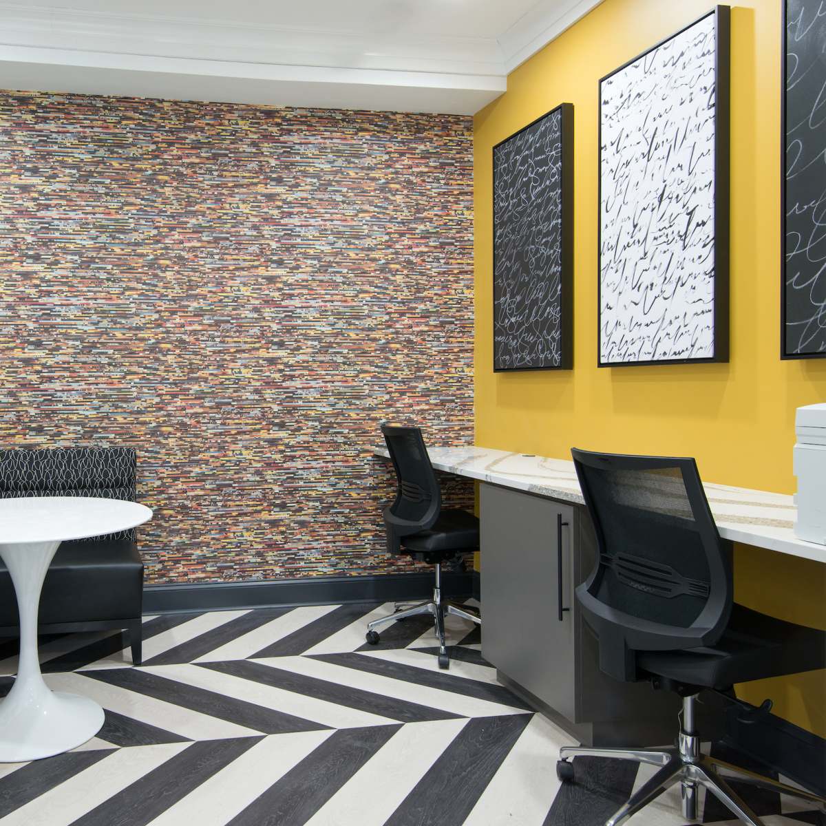 A modern lounge area with a patterned wall, a round white table, black seating, and two workstations against a yellow wall.