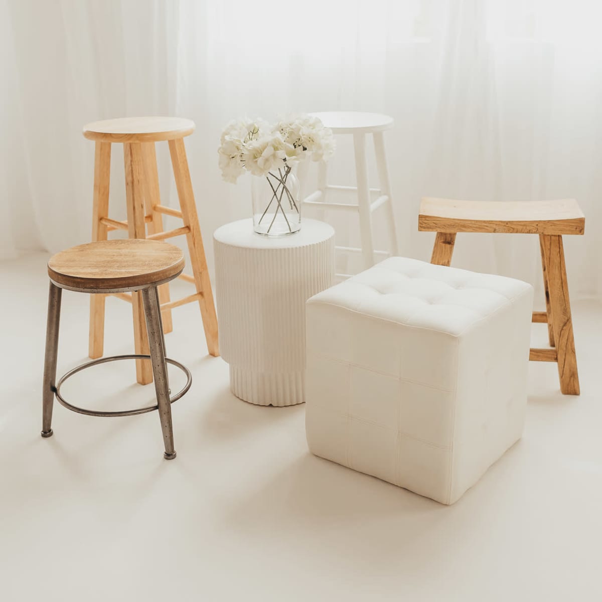 A small arrangement of five stools and a round white table is placed on a light-colored floor, with a vase of flowers on the table.