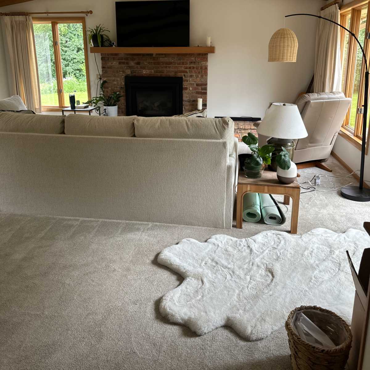 A cozy living room featuring a beige couch, a brick fireplace with a television above it, and large windows allowing natural light to enter.