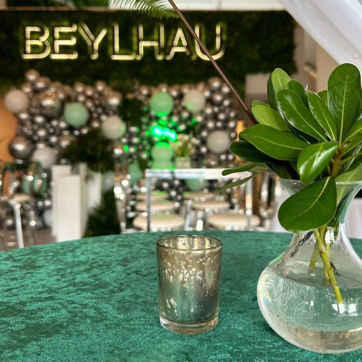A table with a green tablecloth features a small vase of leaves and a decorative candle holder in front of a backdrop with the name "BEYLHAU" and a balloon arrangement.