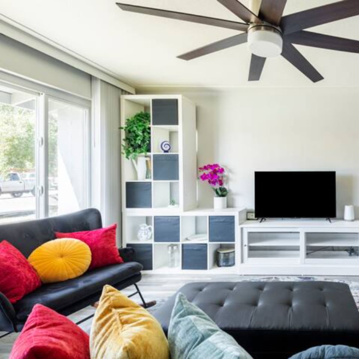 The living room features a modern design with a black ceiling fan, a dark sofa, colorful cushions, a sleek entertainment unit, and large windows allowing natural light.