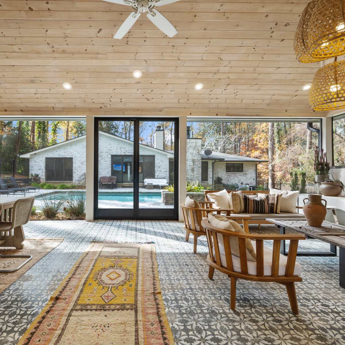 A bright, open living space with modern decor, featuring a wooden ceiling, large glass doors leading to a pool area, and a mix of natural materials in the furniture and decor.