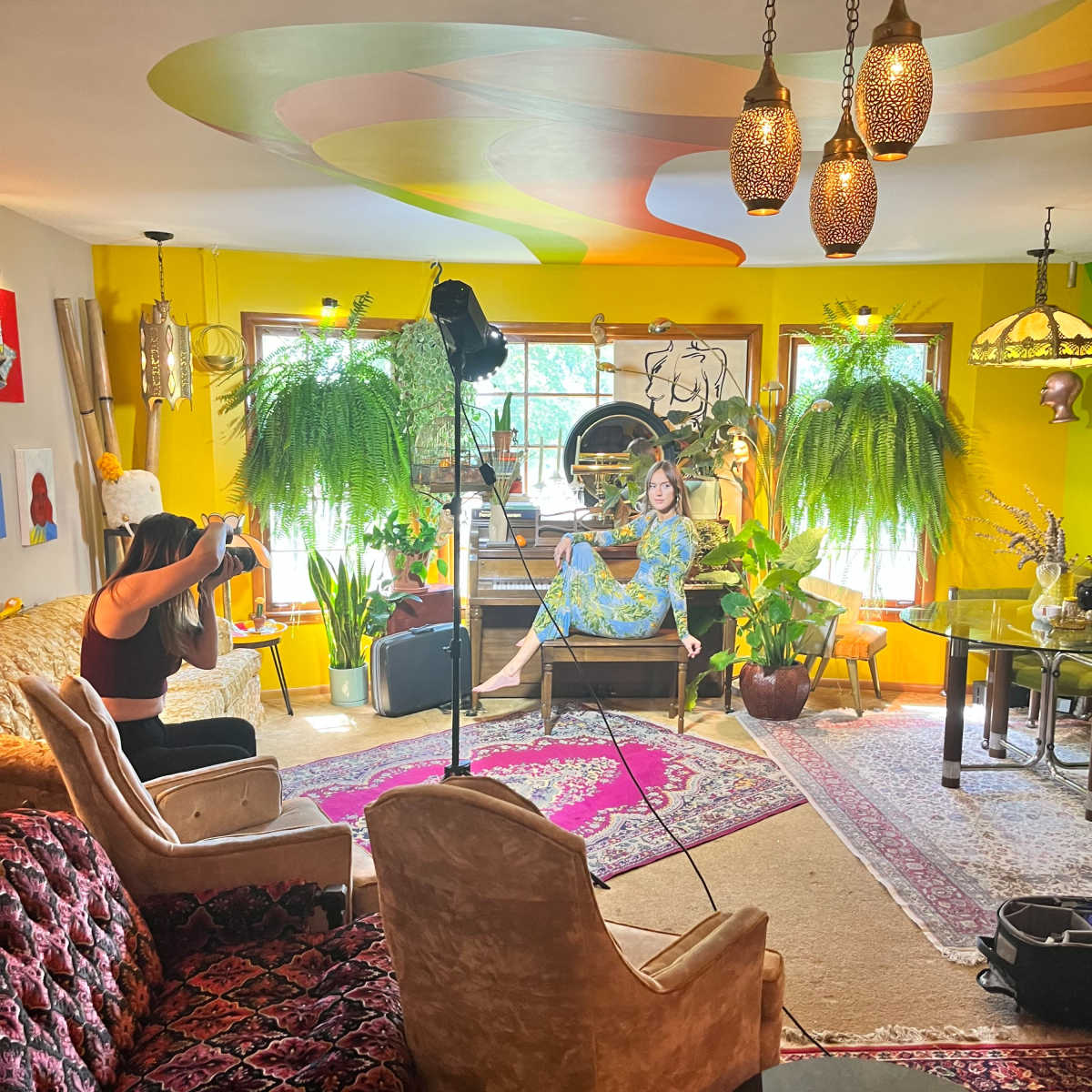 A person is photographing a model sitting in a brightly colored, plants-filled living room with eclectic decor.