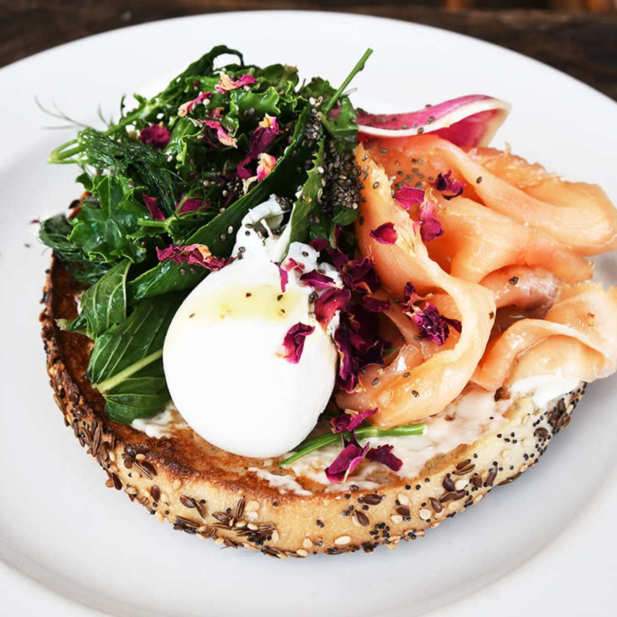 A toasted bagel topped with smoked salmon, a poached egg, greens, and a garnish of rose petals.