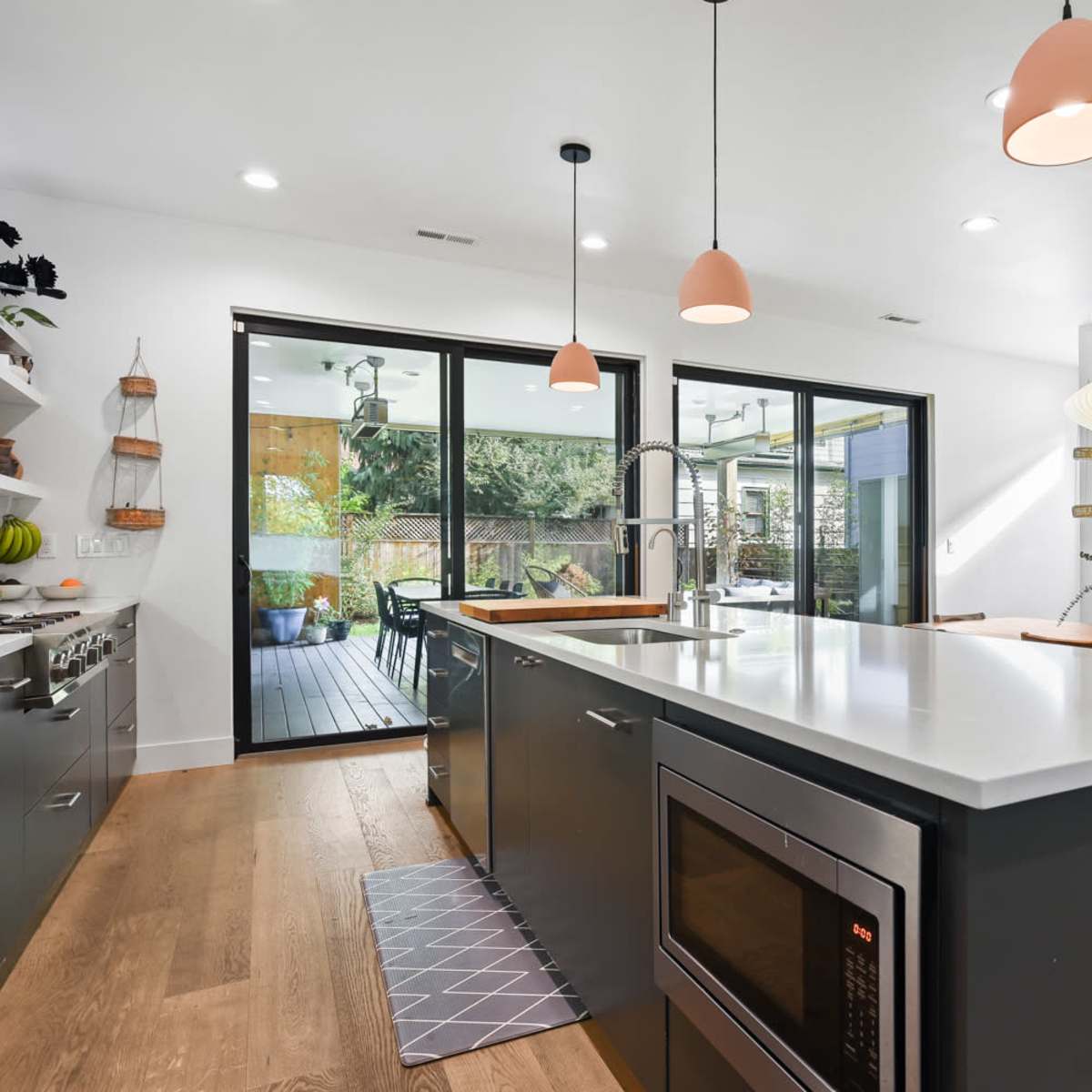 A modern kitchen features an island with a sink and microwave, light fixtures above, and large glass doors opening to an outdoor space.