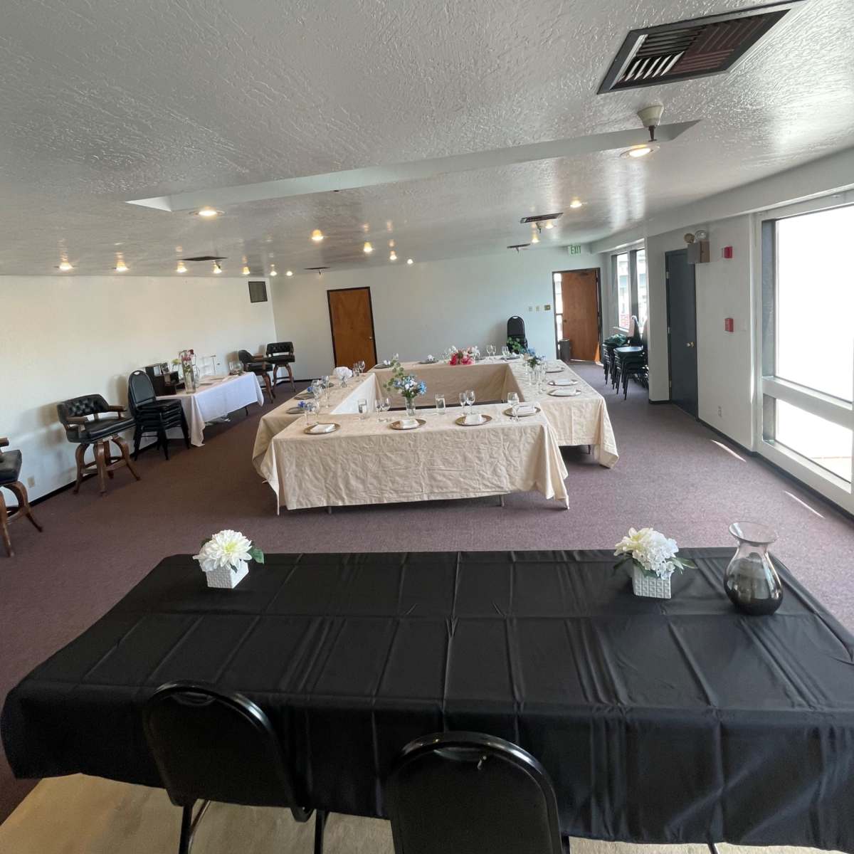 A spacious event room with two tables set for a gathering, one clothed in black and the other in beige, surrounded by chairs and a serving area.