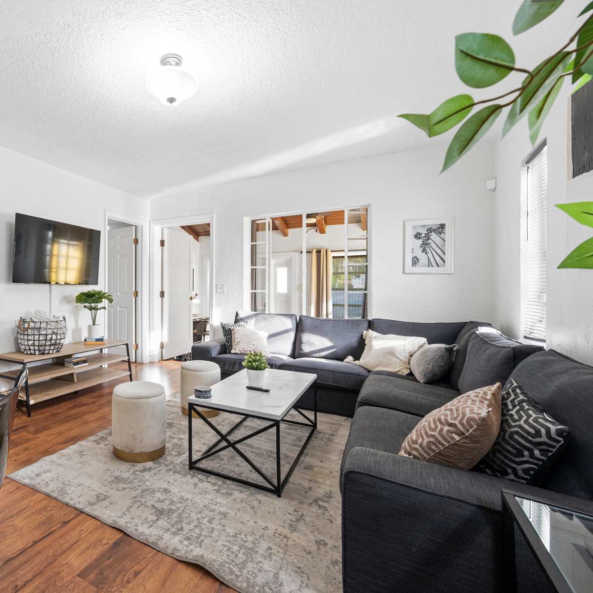 A modern living room with a dark sectional sofa, a coffee table in the center, and a dining area featuring a glass table and chairs, all complemented by bright natural light.