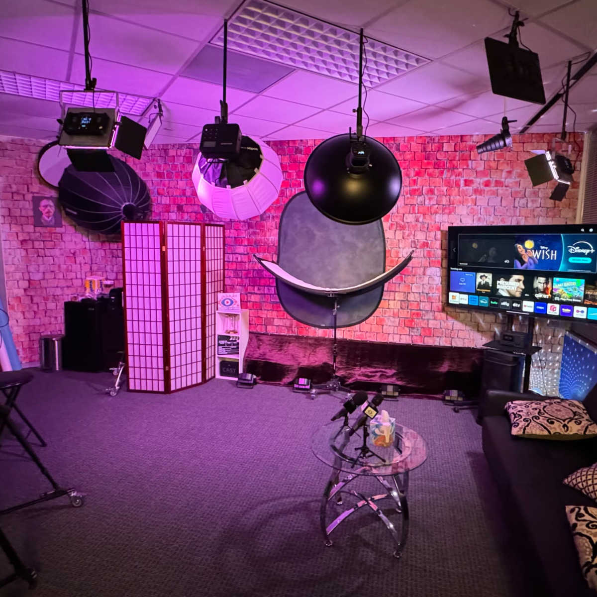 A studio setup with a black sofa, a glass coffee table, a television screen, and various lighting equipment against a brick wall backdrop.