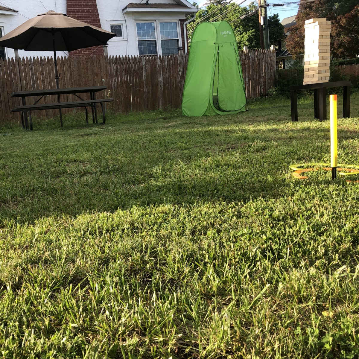 A grassy backyard featuring a picnic table with an umbrella, a green tent, and a game setup on a black table.