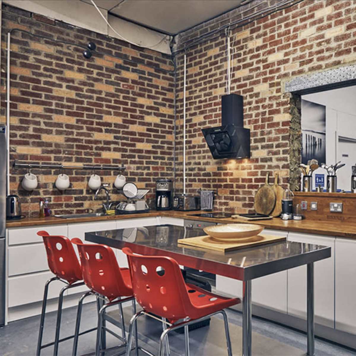The kitchen features a brick wall, stainless steel countertops, and red plastic chairs around a rectangular table.