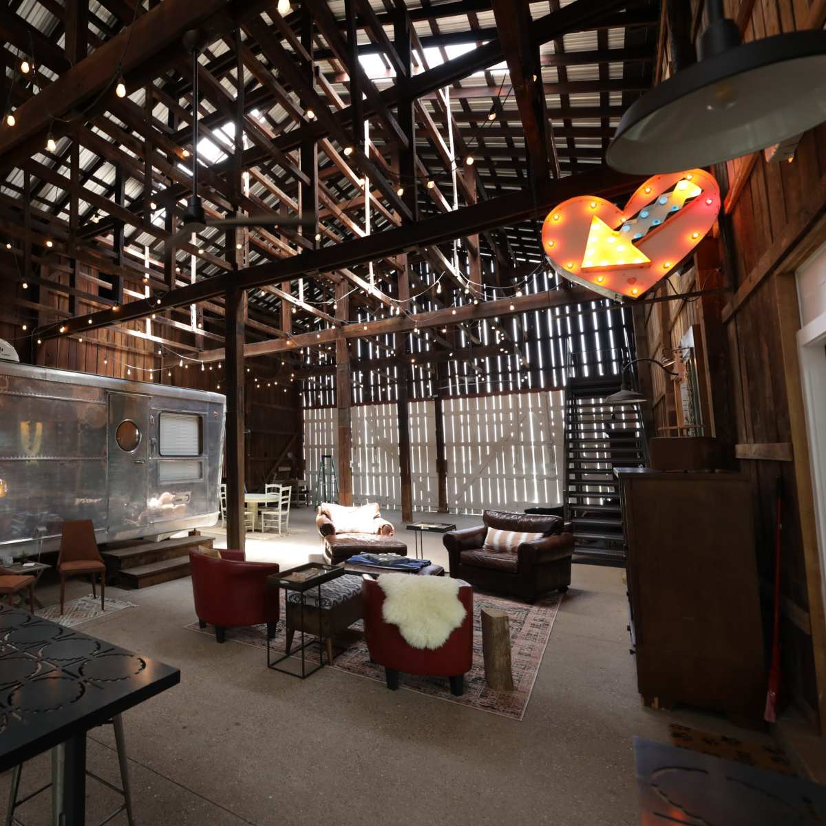 The interior of a barn, featuring a shiny metal trailer, rustic seating arrangements, and string lights illuminating the wooden beams.
