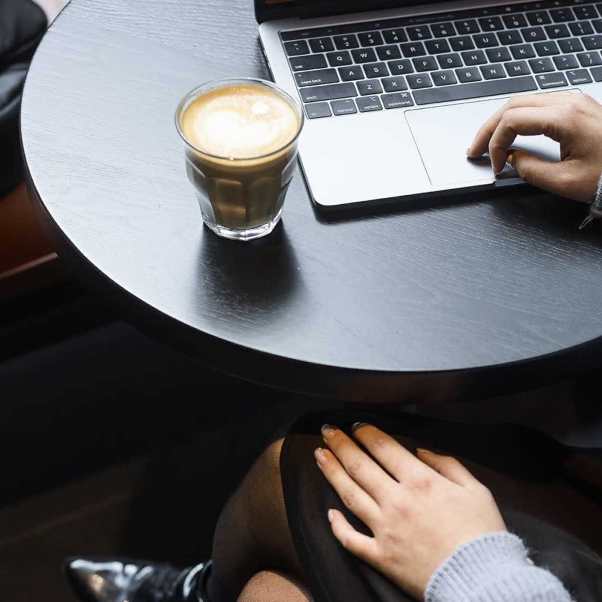 A person is seated at a black table, using a laptop while a glass of coffee sits beside them.