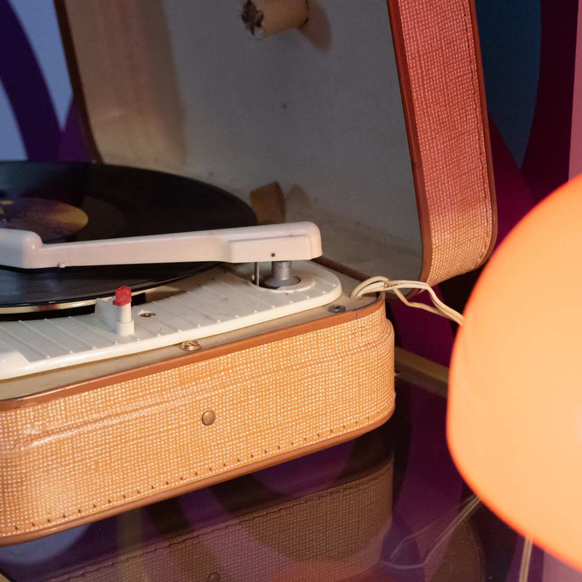 A vintage record player is positioned next to an orange lampshade on a colorful patterned background.