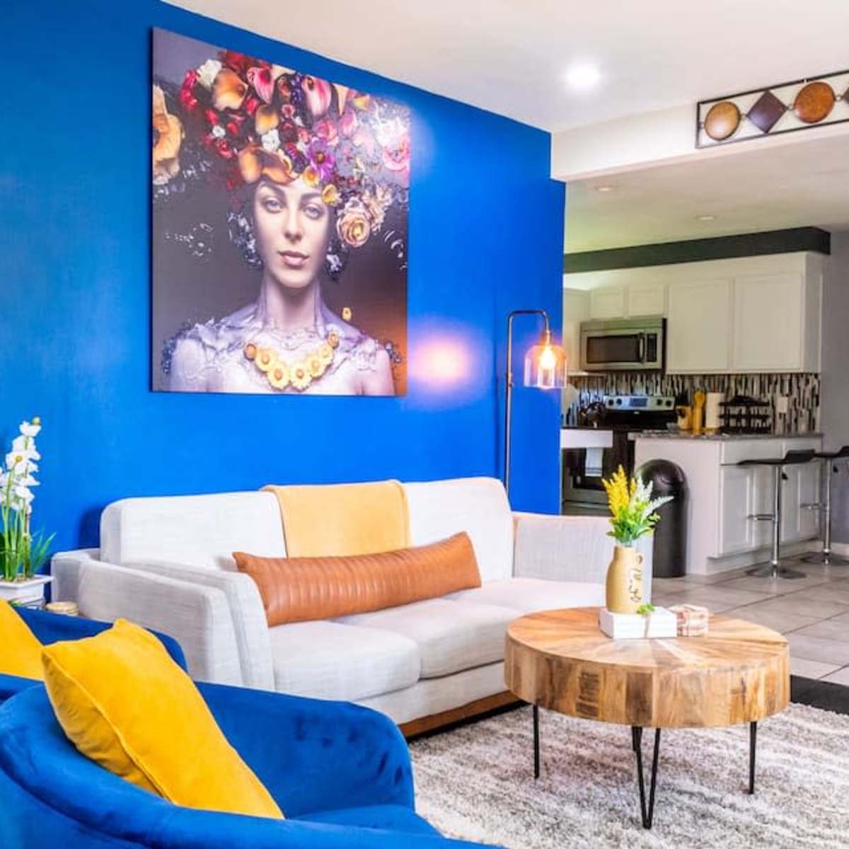A modern living room with blue walls, a large artwork featuring a woman with flowers in her hair, a gray sofa, yellow accent chairs, and a round wooden coffee table.