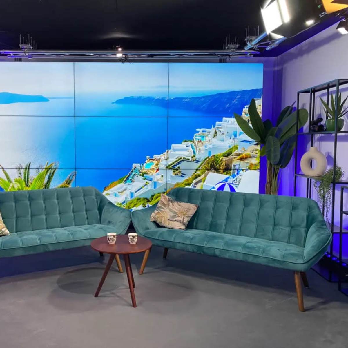 A modern television studio setup featuring two teal sofas and a small round table, with a large screen displaying a coastal view in the background.