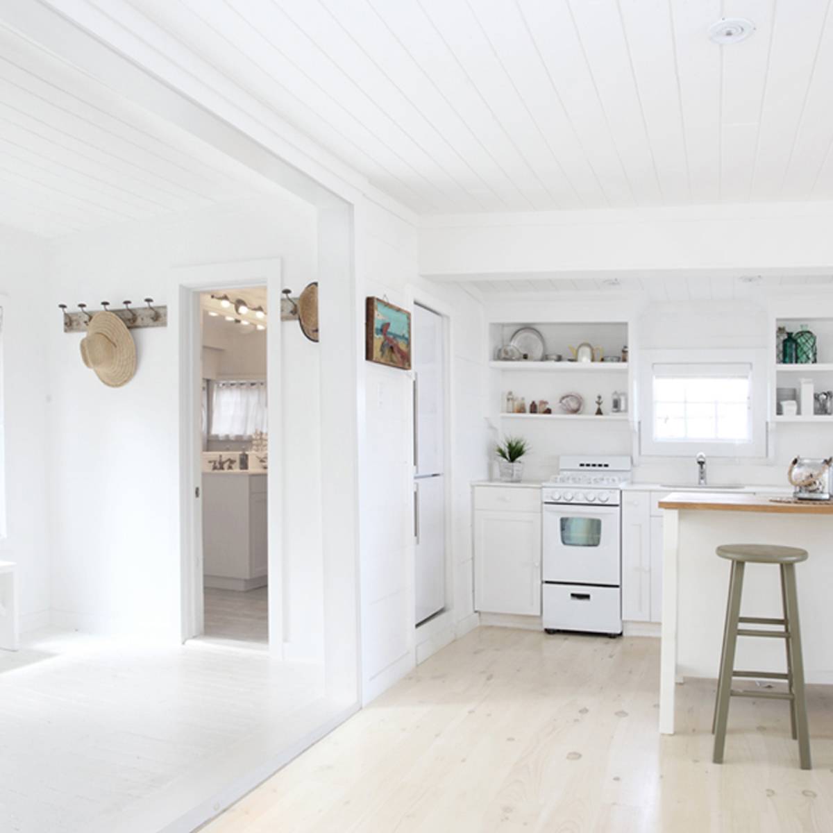 A bright, open kitchen and living area with white walls, wooden floors, and a small island with two stools.