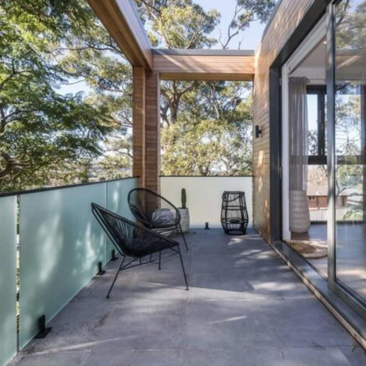 A spacious balcony with a wooden overhang, a concrete floor, and large glass doors leading into a modern interior, surrounded by trees.
