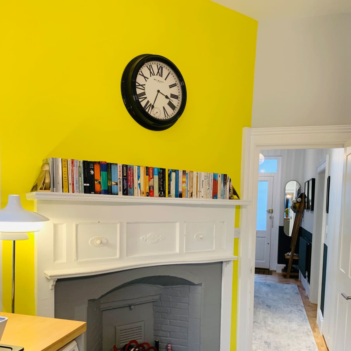 A brightly colored yellow wall with a clock above a white mantelpiece lined with books, and a view into a hallway with a patterned floor.