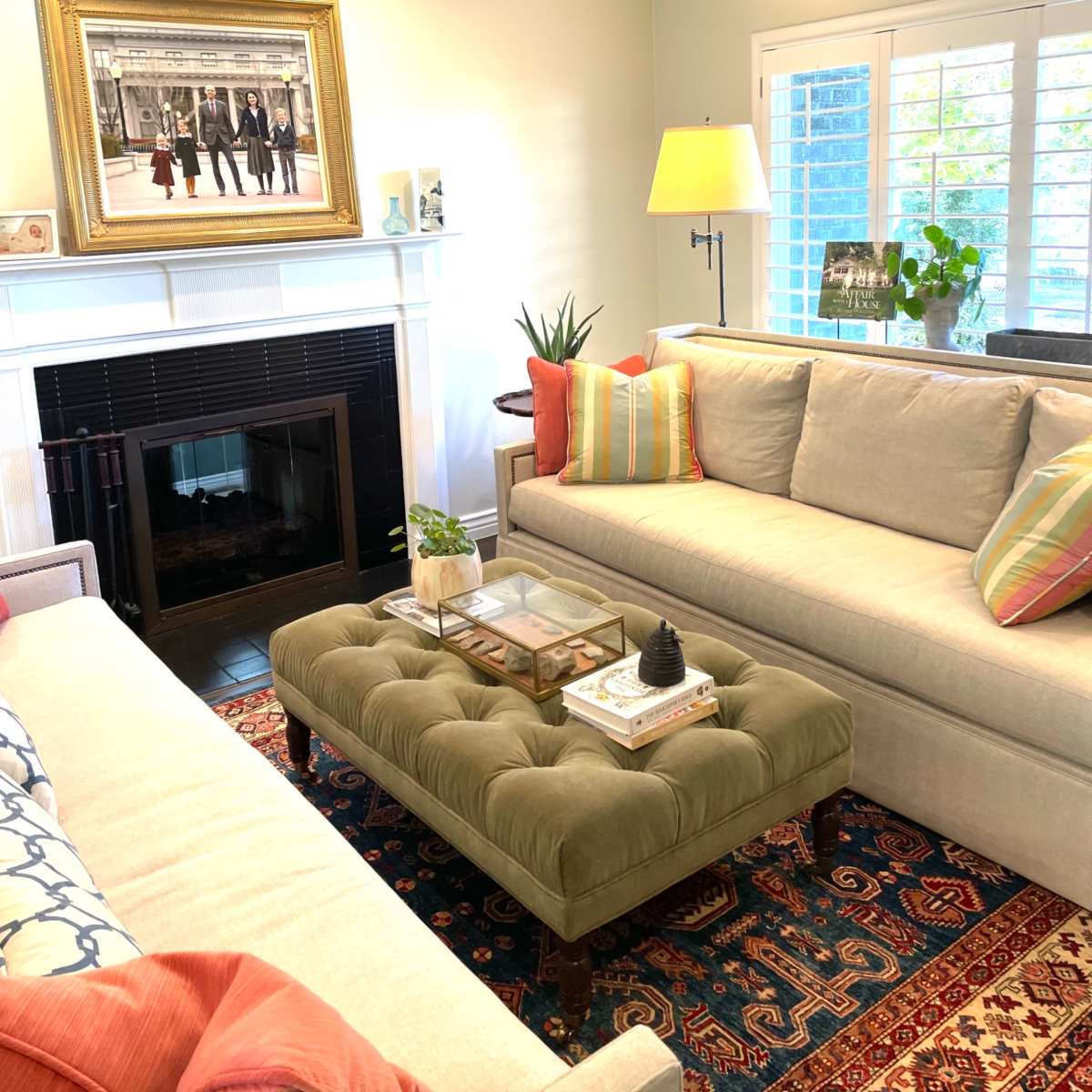 A bright living room features two sofas, a tufted coffee table, a framed photo above a fireplace, and natural light coming through window shutters.