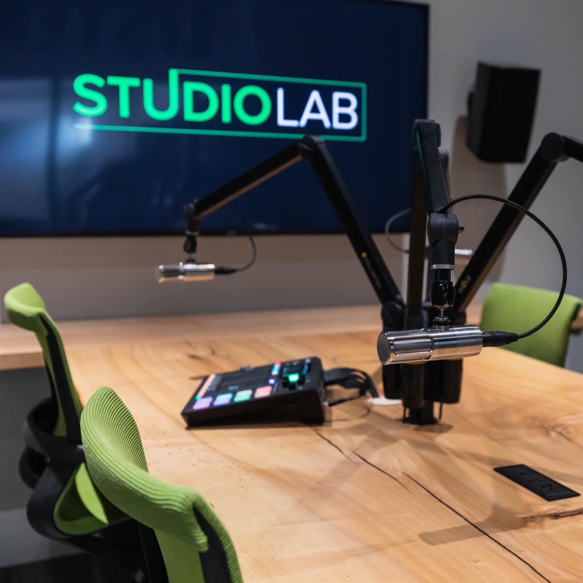 A recording studio setup featuring two adjustable microphones mounted on a wooden table, with a control panel and a large screen displaying "STUDIOLAB" in the background.