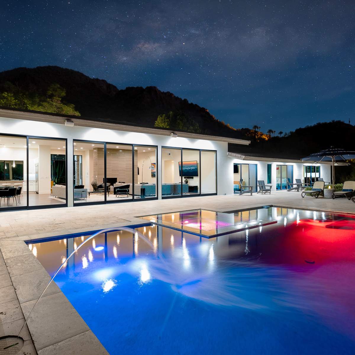 A modern home with a swimming pool illuminated in different colors under a starry night sky.