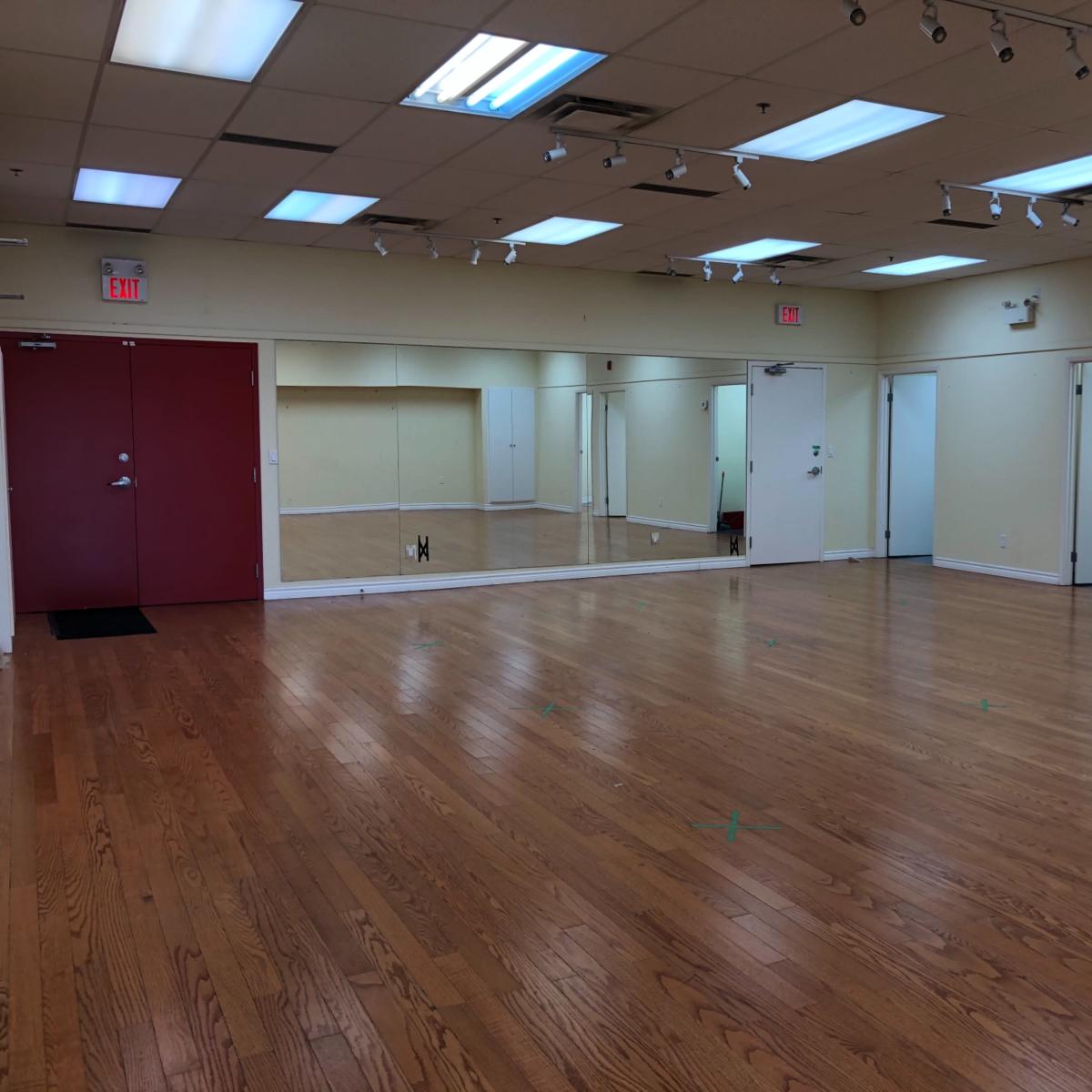 A spacious, empty room with hardwood floors, bright overhead lights, and several mirrors on one wall.