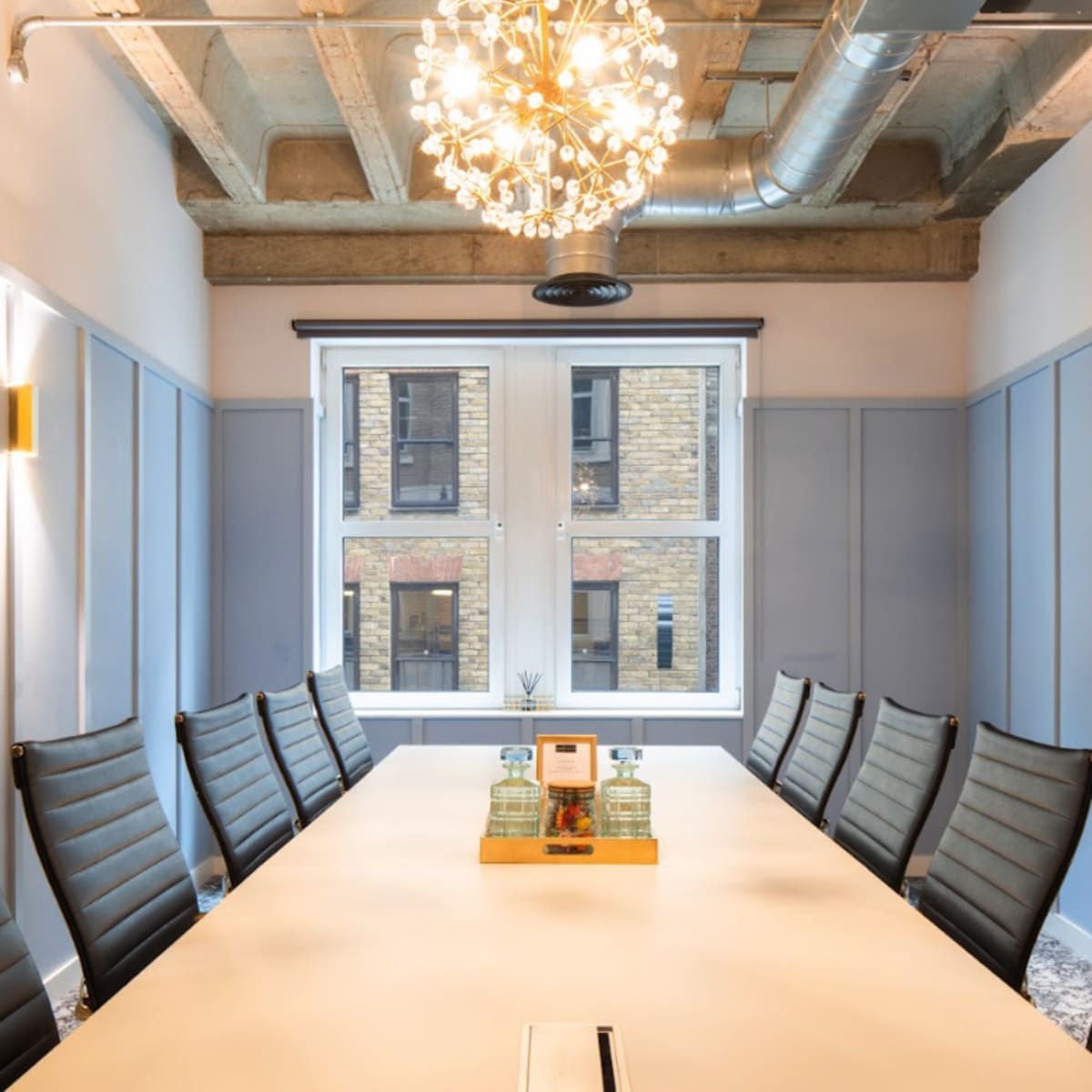 A modern conference room with a long table, eight ergonomic chairs, and a decorative chandelier above, illuminated by natural light from a large window.