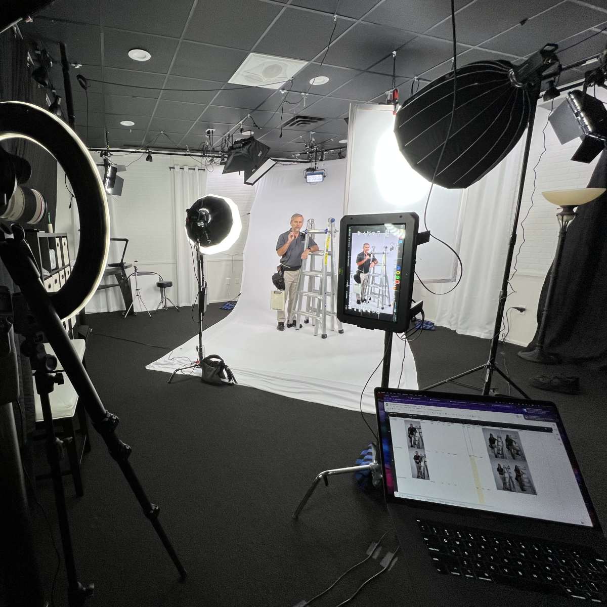 A photo studio setup features a seated subject under bright lights, with camera equipment, a laptop displaying images, and various lighting tools arranged around a white backdrop.