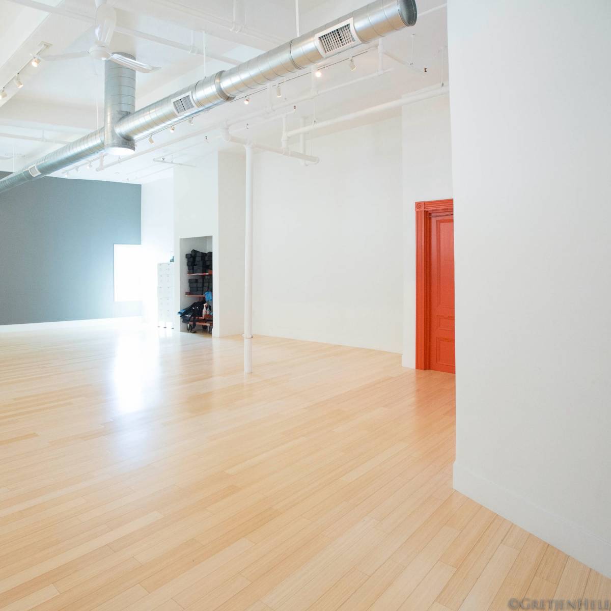 A spacious, empty room with light wood flooring, a gray accent wall, and a bright orange door.
