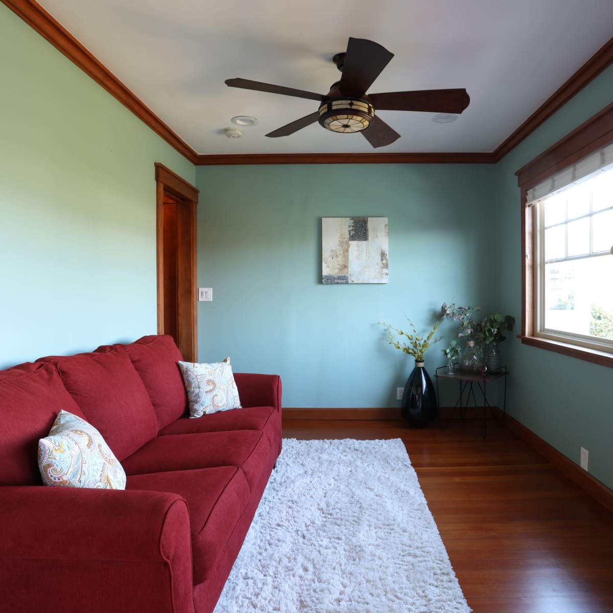 A bright room with a red sofa, a light rug on hardwood flooring, and a window with wooden trim.