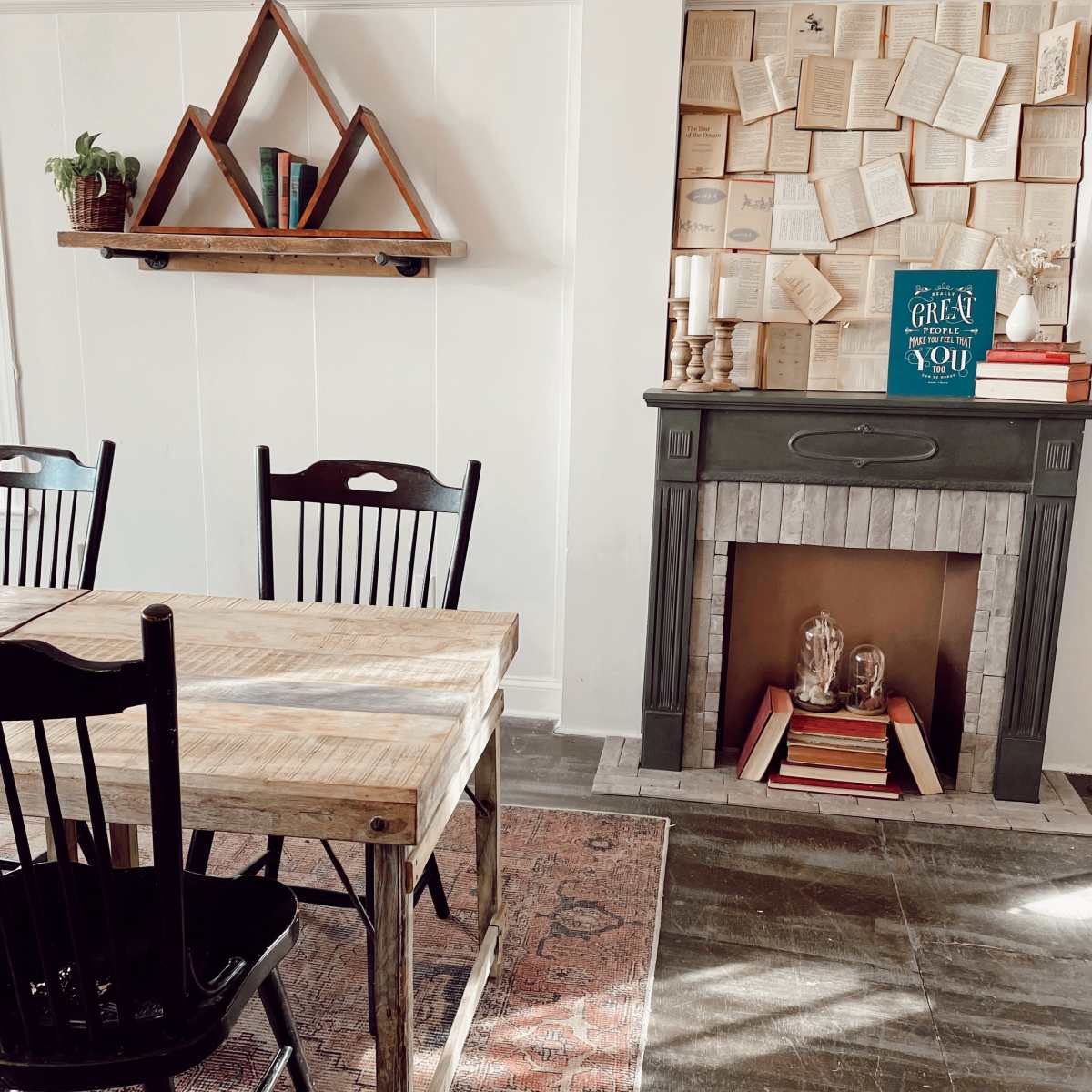 A dining area features a wooden table with black chairs, a shelf displaying plants and decorative items, and a fireplace surrounded by books and candles.