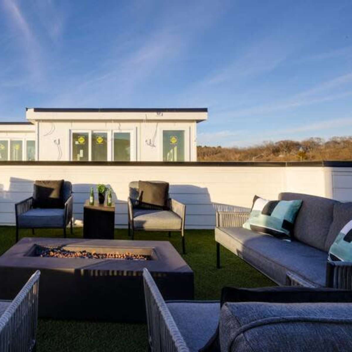A rooftop patio features seating arranged around a central fire pit with green landscape accents and modern furnishings.