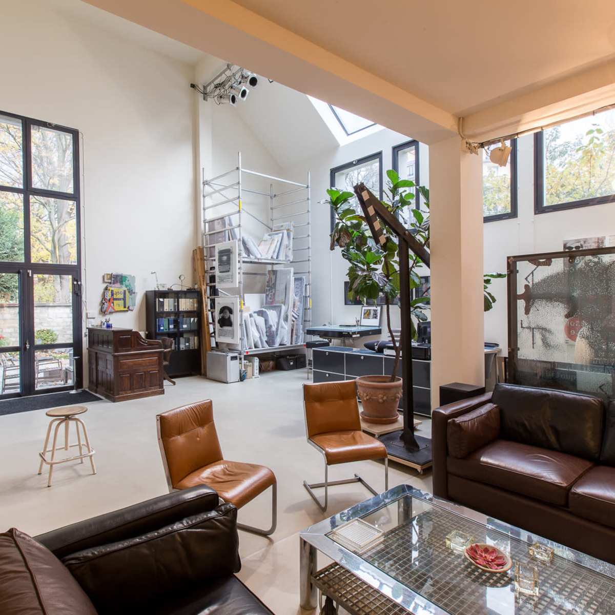 An open-concept living area featuring leather seating, a glass coffee table, large windows, and a workspace with a plant in the background.