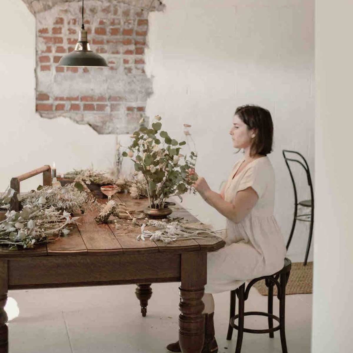 A woman sits at a wooden table adorned with dried flowers, while a brick wall serves as the backdrop.