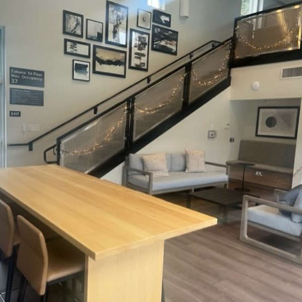 A modern lobby area with a wooden table, chairs, and a staircase adorned with framed pictures and decorative lights.